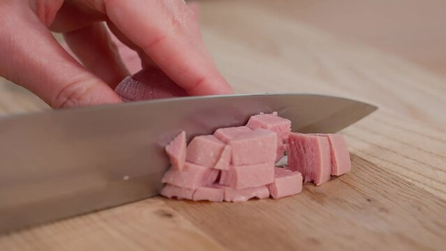 The ham is cut into cubes with a sharp knife on a wooden board. Ham filling for breakfast. Appetizers with ham. Close-up, macro, side view