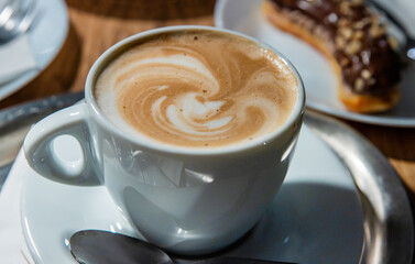 Cup of coffee latte with cream foam served on the white plate with spoon. Few plates on a wooden table in coffee shop. Mug of milk coffee, cappuccino, eclair with chocolate glaze. Selective focus