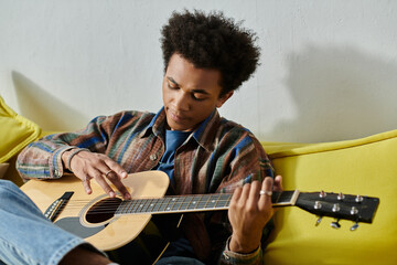 A young African American man sitting on a couch, playing an acoustic guitar while talking on the...