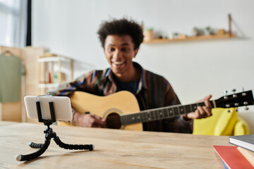A young African American male blogger playing guitar on a tripod while speaking into a cell phone...