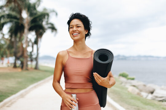 Smiling woman with yoga mat walking along seaside promenade in sportswear outdoors