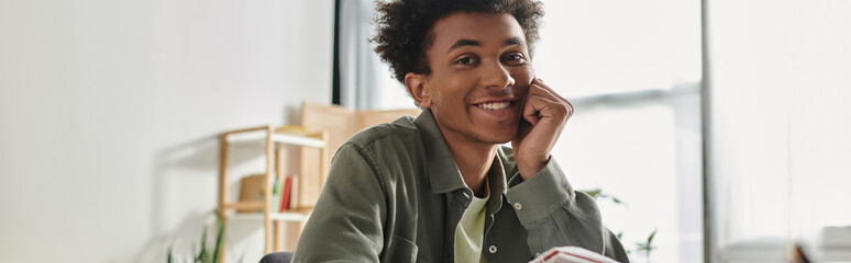 A young man, African American, sitting at a desk with a book, studying online at home.