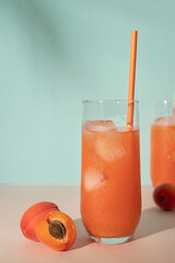Juicy iced apricot drink in glass with straw, fruits on pink table, cyan background with soft floral sunlight shadows, summer refreshing beverage, retro style