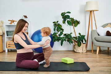 A beautiful young mother sitting on a yoga mat, cradling her baby with the guidance of a coach at...