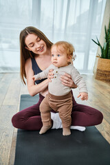 A young mother gently holds her baby on a yoga mat, guided by a coach at parents courses in the...