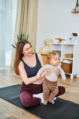 A young, beautiful mother sits on a yoga mat, peacefully cradling her baby with the guidance of her...