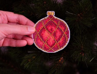 A woman hand holding a red cross stitched Christmas ornament against a Christmas tree background.
