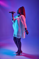 African-American woman wearing elegant white dress singing into microphone in neon light against...