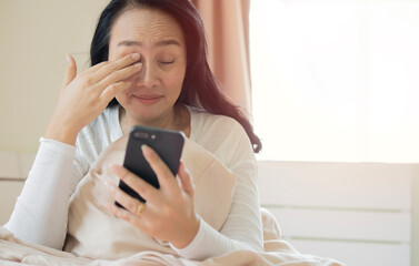 Happy mature woman in pajamas rubbing eyes and using mobile phone on bed at bedroom.