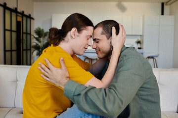 A young gay couple in casual clothing embraces on a white couch in their modern apartment.