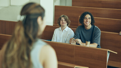 Focus on two boys, classmates sitting in university auditorium and passing exam to female teacher. Concept of education, youth, workshop, knowledge