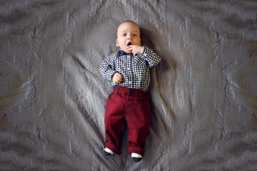A baby in a plaid shirt and maroon pants lies on the bed