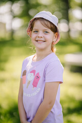 Valmiera, Latvia - June 4, 2024 - Smiling young girl wearing a purple shirt with ice cream graphics and a sunhat, standing outdoors on a sunny day.