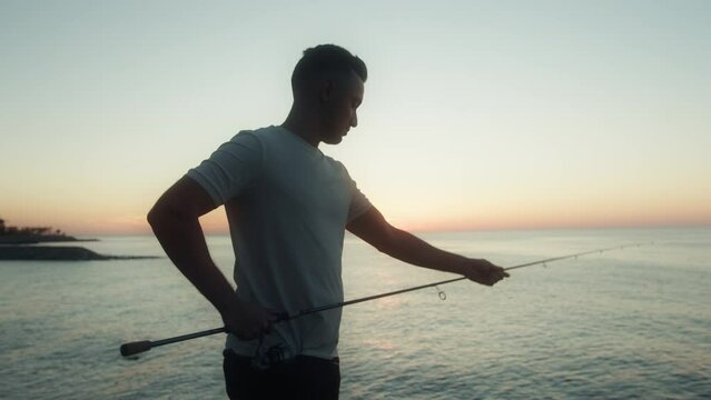 Young man with a fishing rod stands on the shore and enjoying a beautiful sunset. Fisherman fishing on the ocean and enjoying nature