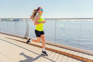 Woman in a bright yellow tank top jogs along a waterfront boardwalk on a sunny day.