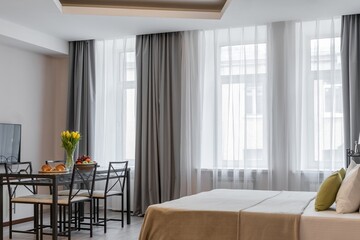 The design of a modern bedroom with a large bed and large panoramic windows. A glass table with yellow flowers in a vase, fruits and croissants. Large chairs.