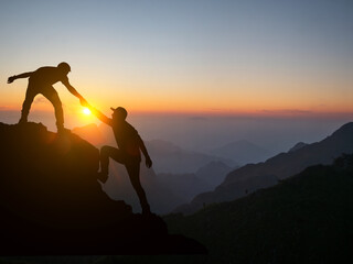 Silhouetted image of two climbers helping each other atop a mountain peak at sunrise, showcasing...