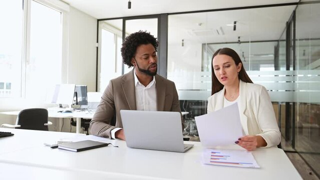 Two business colleagues engaging in a constructive discussion with a focus on paperwork, signifying teamwork and cooperation in a corporate environment, using laptop and solving tasks together