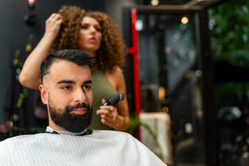 Portrait of a man in a barbershop with a female barber in the background, crafting a stylish look.