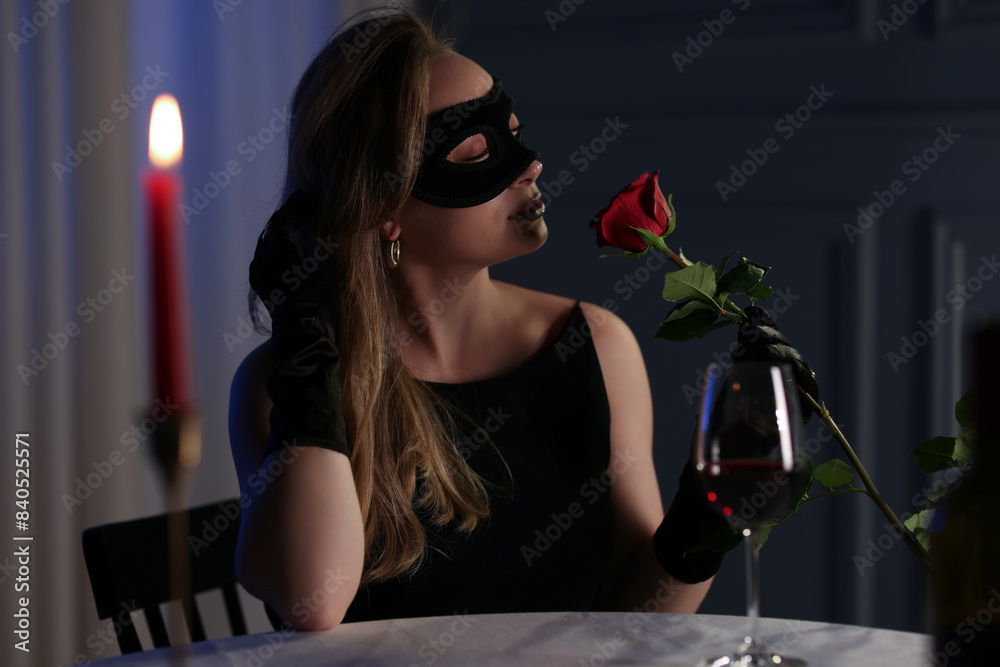 Wall mural elegant woman in black eye mask with rose at table indoors in evening