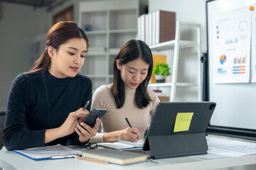 Two women are sitting at a desk with a laptop and a cell phone