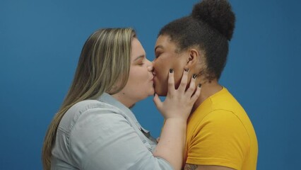 Sweet and tender kiss between two women in love shown of affection on isolated blue background studio. Free love to all sexual orientation. Gay young relationship.
