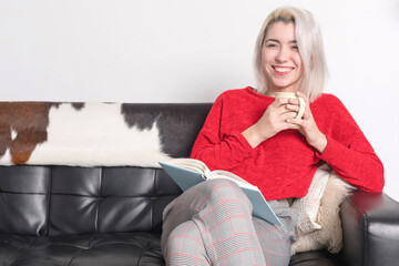 Smiling woman in red sweater with a mug and book on a couch.