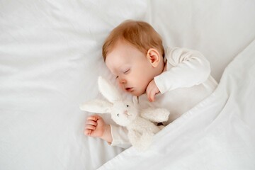 A baby boy sleeps under a blanket in a white bodysuit on the bed with his hand folded under his cheek and holding a soft toy, a sweet healthy baby sleep at home