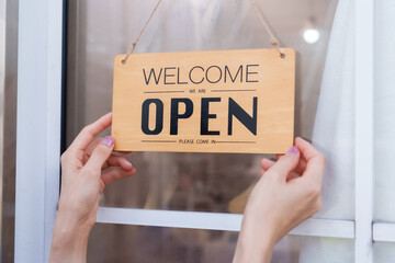 Beautiful Asian girl small business spa owner hanging open sign at door. Wearing blue apron and light blue shirt, ready to welcome customers. Professional, inviting, clean spa environment.