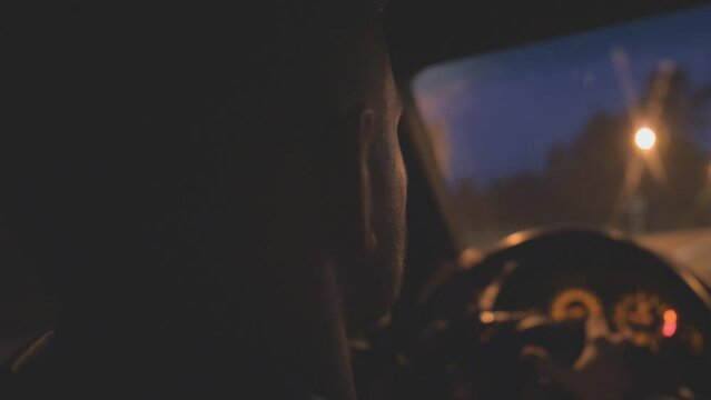 Close up of young man holds hands on steering wheel and drives a modern automobile at evening. Guy rides in auto on highway at night time. Male driver moves in his SUV car on empty road. Slow mo