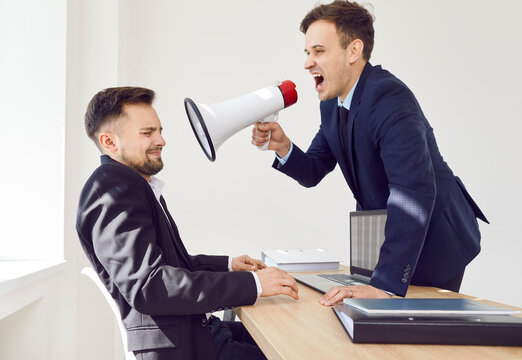Young angry businessman wearing suit shouting with megaphone on a stressed man company employee working on laptop sitting at the desk on workplace in office. Angry boss yelling at his male worker.