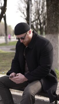 A man is sitting on a bench in the park, using his smartphone outdoors under the trees