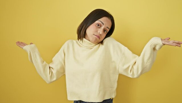 Clueless young hispanic woman stands doubted, her beautiful face alone against a yellow wall, hands and arms raised in confusion, wearing a sweater.