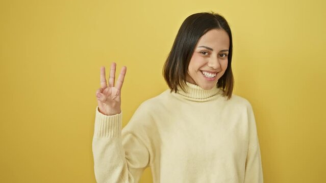 Cheerful young hispanic woman in yellow backdrop, smiling confidently, raising three fingers in a gesture of showing 'number three'. wearing a stylish sweater, a picture of beauty.