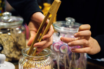 Teenager girl selling candies at the food market. Picking out candy with wooden or bamboo tongs out of the glass jar and putting them into plastic bag. Copy space. Selective focus, Blurred hands