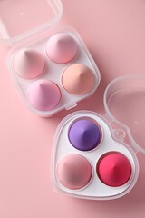 Makeup sponges in plastic containers on pink background, top view