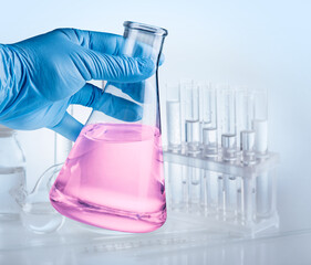 Conical flask in hand in blue glove and laboratory equipment at the background.