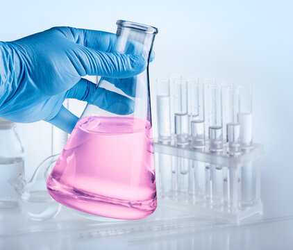 Conical flask in hand in blue glove and laboratory equipment at the background.