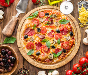 Pizza topped with cherry tomatoes, salami and other ingredients is on wooden table surrounded by ingredients. Top view.