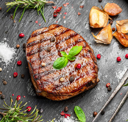 Grilled delicious ribeye steak and some seasonings and herbs on black slate serving plate. Flat lay.