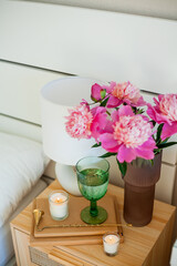 Bouquet of peonies in glass pink vase, lamp, candles on wooden stand. Bedroom. Spring flowers.