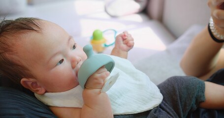 A close-up of a baby lying down and chewing on a blue teething toy looks content and focused....