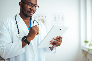 A focused black male doctor reviews medical information using a digital tablet, dressed in a white lab coat with a stethoscope, standing in a well-lit doctor's office.