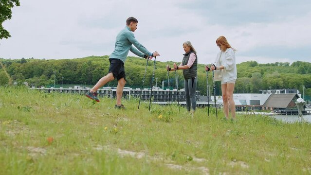 Man trainer shows how to use Scandinavian trekking sticks. People hold nordic poles in green forest park with lake. Cloudy weather