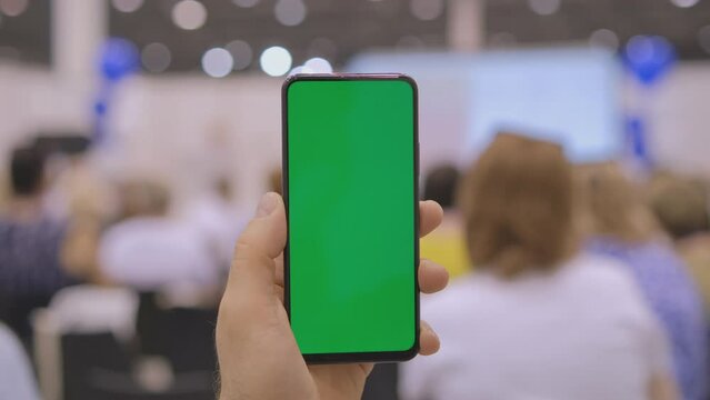 Person showcasing green screen smartphone at conference with focused audience in blurred background
