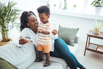 A young black mother engages with her son in playful conversation, comfortably seated on a sofa in a well-lit living room, creating a nurturing family moment.
