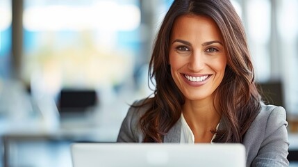 A woman in a business suit sits in an office, smiling confidently as she works on her laptop