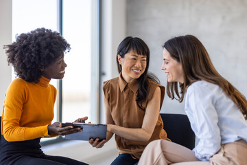 Three women of varied ethnic backgrounds in professional attire discuss work, sharing insights over a clipboard in a modern office setting.