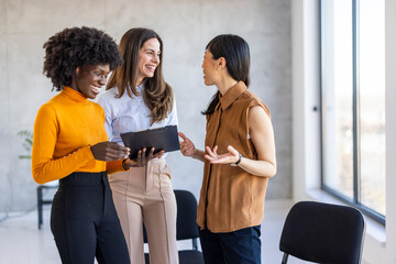 A diverse trio of professional women engage in a dynamic conversation in a bright office, reviewing...
