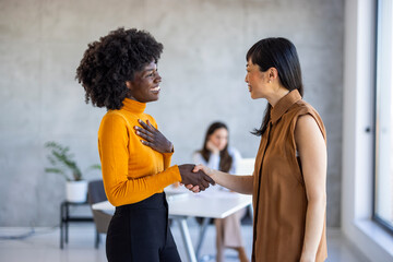 A cheerful Black businesswoman in a vibrant orange top clasps hands with an Asian colleague wearing a sleeveless brown blouse, exemplifying professional collaboration within a sleek